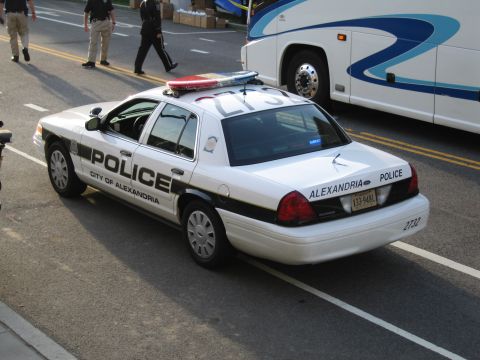 1991 Ford LTD Crown Victoria - Rear three-quarter view of a black-and-white City of Alexandria Police sedan parked beside a tour bus with officers walking.