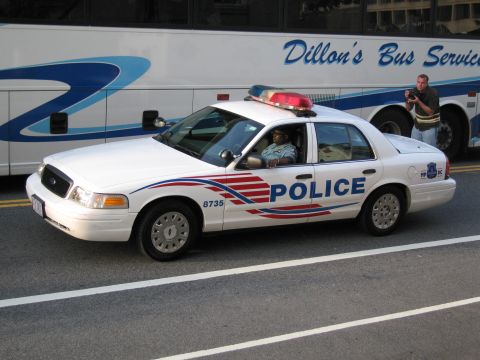 1991 Ford LTD Crown Victoria - white Metropolitan Police sedan lettered POLICE sits with an officer inside next to a Dillon's Bus Service motor coach.