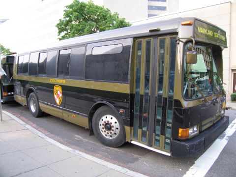 1991 Ford LTD Crown Victoria - Front three-quarter view of a black and gold Maryland State Police bus with a state crest, parked curbside in a city.