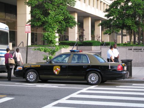 1991 Ford LTD Crown Victoria - black and tan Maryland State Trooper sedan parked curbside near pedestrians, a transit bus, and a courthouse.