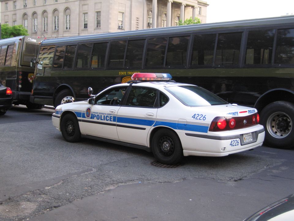 White and blue county Police Impala sedan beside a dark bus