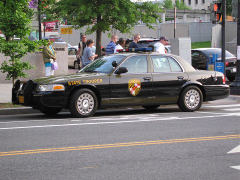 1991 Ford LTD Crown Victoria - black and tan Maryland State Trooper sedan parked on a street as several people stand on the sidewalk behind it.