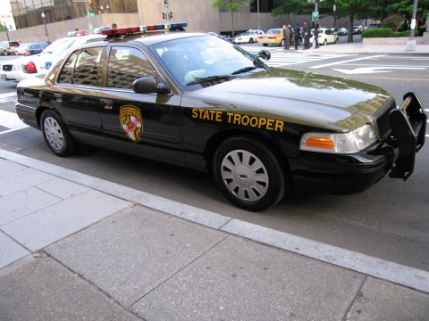 1991 Ford LTD Crown Victoria - black and tan Maryland State Trooper Ford sedan with its front door open, parked curbside near an intersection.