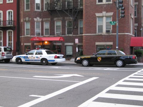 1991 Ford LTD Crown Victoria - white Police sedan and a black and tan State Trooper sedan parked along a brick city street by apartment buildings.