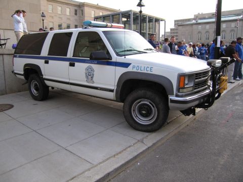 1991 Ford LTD Crown Victoria - white and blue Police Chevrolet Suburban SUV with a light bar parked on a paved plaza near a crowd of people.