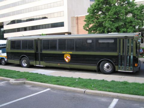 1991 Ford LTD Crown Victoria - Side view of an olive and black Maryland State Trooper bus with a state crest parked in a lot beside an office building.