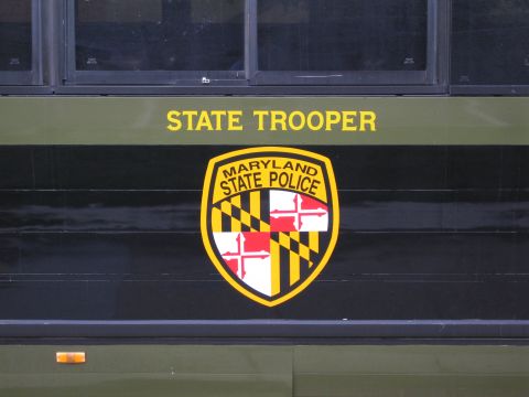 1991 Ford LTD Crown Victoria - Close-up of an olive-drab police vehicle's side bearing yellow STATE TROOPER lettering above a Maryland State Police shield crest.