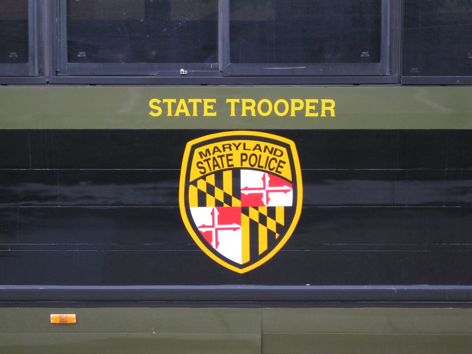 Side of olive-green police vehicle with yellow STATE TROOPER text and Maryland State Police shield emblem