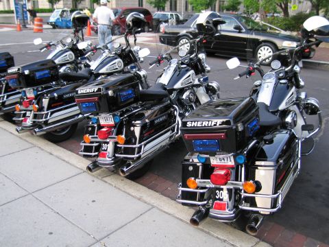 1991 Ford LTD Crown Victoria - Three black police motorcycles marked SHERIFF parked in a row at a city curb, seen from behind with saddlebags and emergency lights.