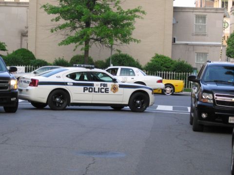 1991 Ford LTD Crown Victoria - black-and-white sedan marked FBI POLICE on its door drives a city street alongside other patrol cars and SUVs.
