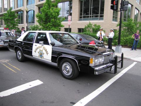 1991 Ford LTD Crown Victoria - black-and-white Ford LTD Crown Victoria marked HIGHWAY PATROL with a star door shield parked along a city curb with officers nearby.