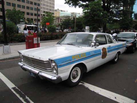 1991 Ford LTD Crown Victoria - white-and-blue vintage Ford police sedan marked POLICE and numbered 601 with a gold badge decal parked at a city curb.