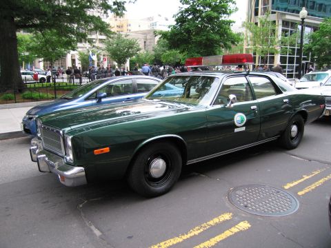 1991 Ford LTD Crown Victoria - dark green vintage Plymouth four-door police sedan with a round green-and-white door decal parked beside a tree-lined city street.