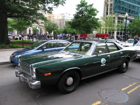 1991 Ford LTD Crown Victoria - dark green vintage Plymouth police sedan with a red roof light bar parked at a city curb next to a blue car.