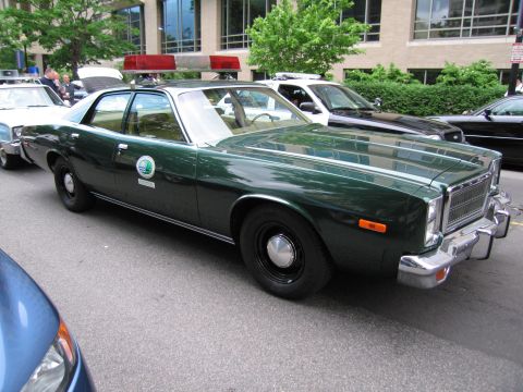 1991 Ford LTD Crown Victoria - dark green vintage Plymouth four-door police sedan with a red roof light bar and circular door emblem parked on a city street.