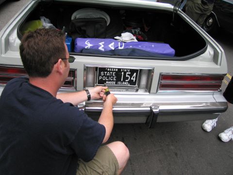 1991 Ford LTD Crown Victoria - man in a dark shirt kneels at an open white hatchback's tail, working on a Rhode Island State Police license plate numbered 154.