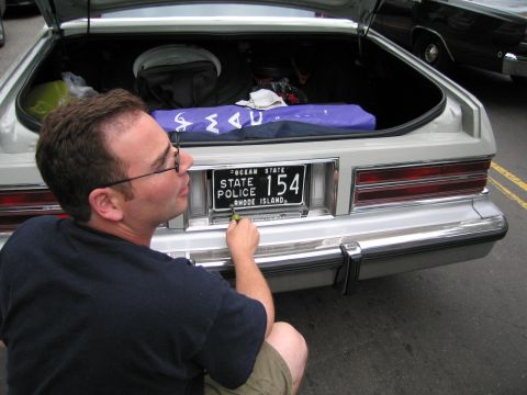 1991 Ford LTD Crown Victoria - bespectacled man kneels at the rear of a white car, holding and adjusting a Rhode Island State Police license plate numbered 154.