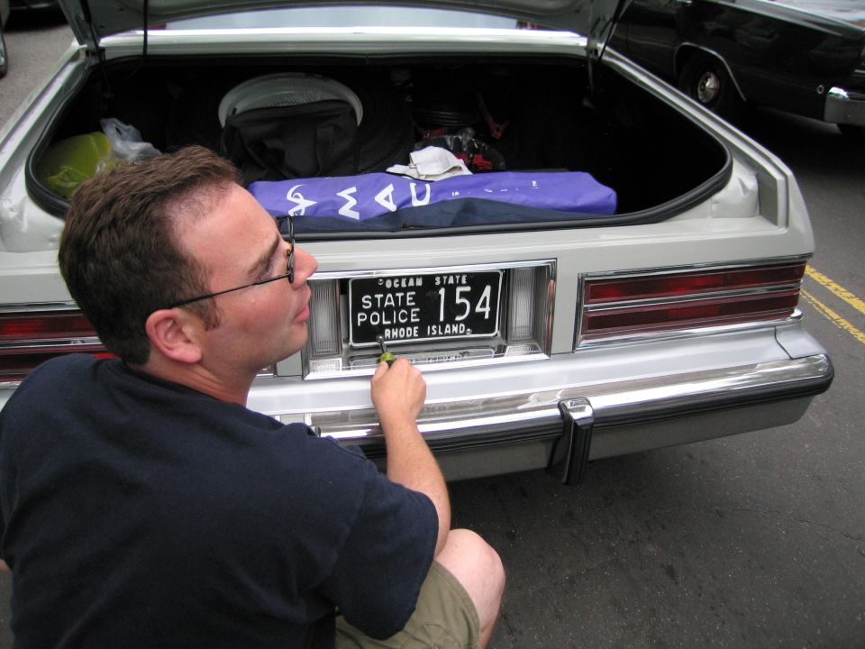 Bespectacled man kneeling behind a white car holding a Rhode Island State Police 154 plate