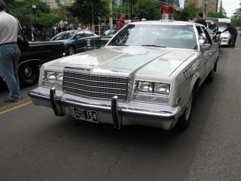 1991 Ford LTD Crown Victoria - Front view of a large silver Chrysler sedan with a roof light and a Rhode Island State Police plate numbered 154, parked among other cars.