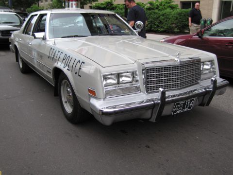 1991 Ford LTD Crown Victoria - white Chrysler full-size sedan lettered STATE POLICE with a roof light and a 154 license plate parked along a curb near a maroon car.