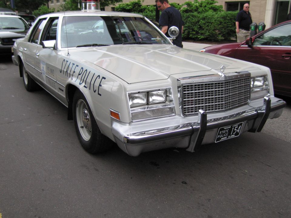 White Chrysler sedan lettered STATE POLICE with roof light and 154 plate parked at curb