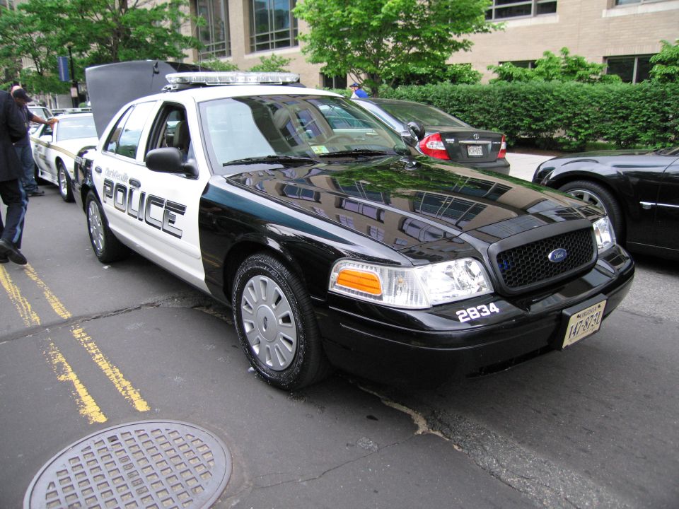 Black-and-white Ford Crown Victoria police car numbered 2834 with roof light bar at curb