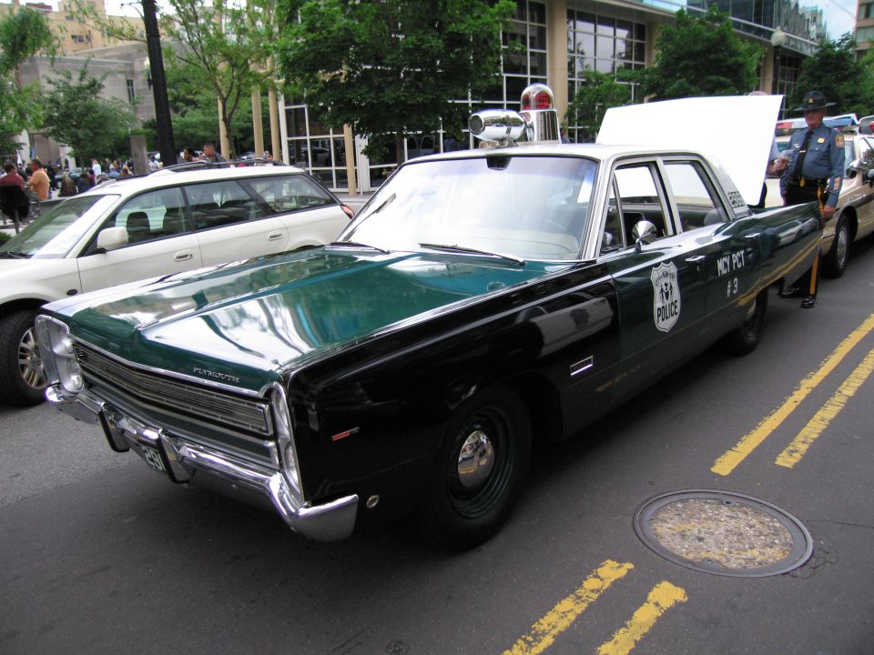 Green-and-black vintage Plymouth NYPD police sedan with roof light bar on a city street