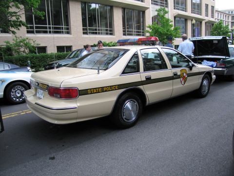 1991 Ford LTD Crown Victoria - tan State Police sedan with green stripe and shield decal, viewed from the rear quarter on a city street.