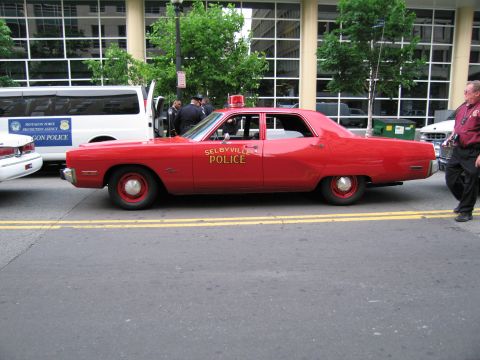 1991 Ford LTD Crown Victoria - Driver-side profile of a red Selbyville Police sedan with red roof light, parked at a curb near a van.