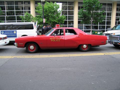 1991 Ford LTD Crown Victoria - Side view of a red Selbyville Police sedan with a red roof beacon, parked at a curb next to a marked van.