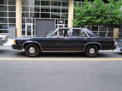 1991 Ford LTD Crown Victoria - dark blue full-size four-door sedan with gold pinstriping and wire wheel covers, parked in profile at a city curb.