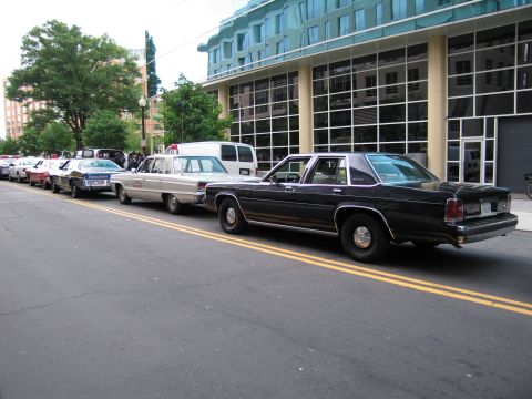 1991 Ford LTD Crown Victoria - line of vintage full-size sedans, including a black one in front, parked along a yellow-lined street beside a curved glass building.