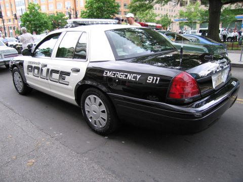 1991 Ford LTD Crown Victoria - Rear quarter view of a black-and-white Police sedan marked Emergency 911, parked along a downtown street.