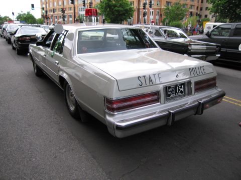 1991 Ford LTD Crown Victoria - Rear quarter view of a silver State Police full-size sedan parked among other cars near a city intersection.