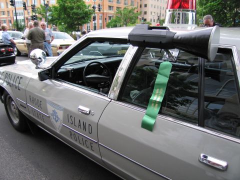 1991 Ford LTD Crown Victoria - Close-up of a Rhode Island State Police sedan showing door lettering, a chrome spotlight, and a green memorial ribbon on the window.