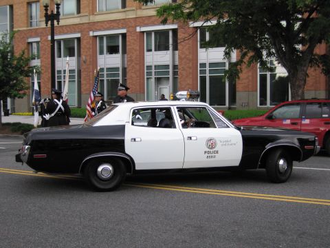 1991 Ford LTD Crown Victoria - black-and-white Police sedan marked 'To protect and to serve' in a street parade, with a uniformed color guard carrying flags.