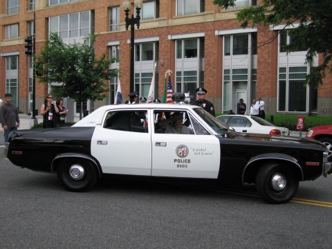 1991 Ford LTD Crown Victoria - black-and-white Police sedan marked 'To protect and to serve' rolling in a parade with officers and flag-bearers around it.