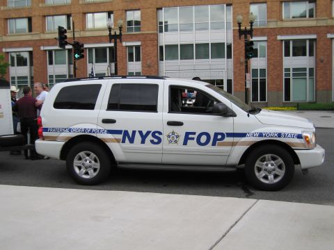 1991 Ford LTD Crown Victoria - white Dodge Durango SUV lettered for the New York State Fraternal Order of Police is parked beside a brick building.