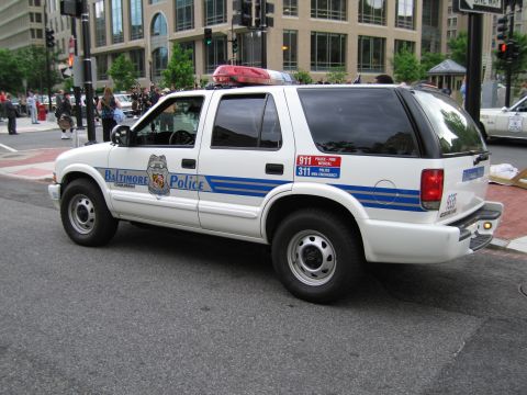1991 Ford LTD Crown Victoria - white Baltimore Police Chevrolet Blazer SUV with blue striping is parked on a downtown city street.