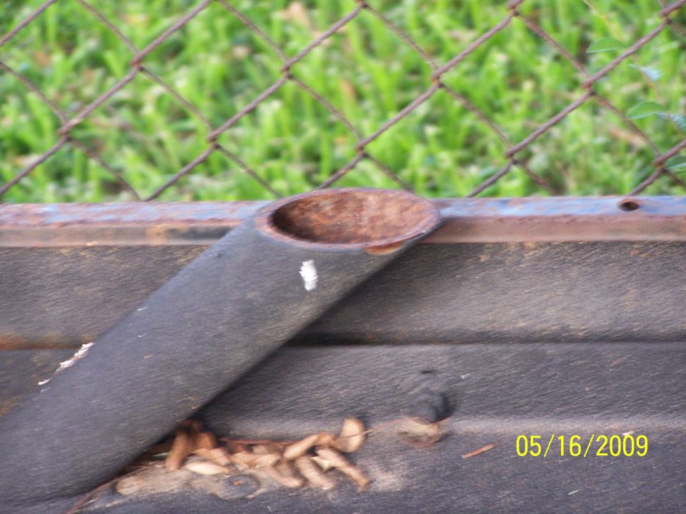Exhaust pipe on a dark textured surface, with a rusty metal bar above and a chain-link fence and grass in the background