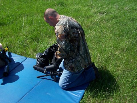 1986 Ford Bronco - A person kneels on a blue mat in a grassy field, surrounded by paintball gear.