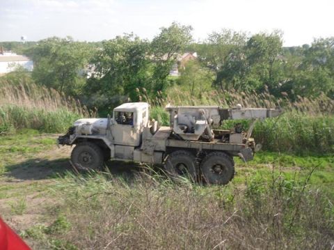 1990 Ford Bronco - A tan military truck equipped with a crane is seen from the side, situated in a grassy, overgrown area.