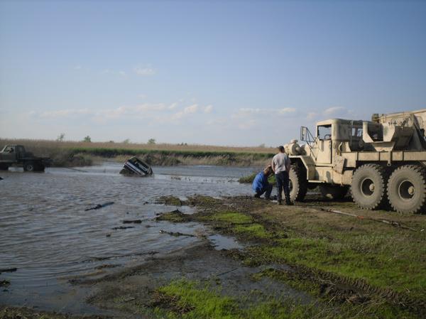 Two trucks are stuck in muddy water, one partially submerged. A large military-style tow truck is on the bank with people nearby