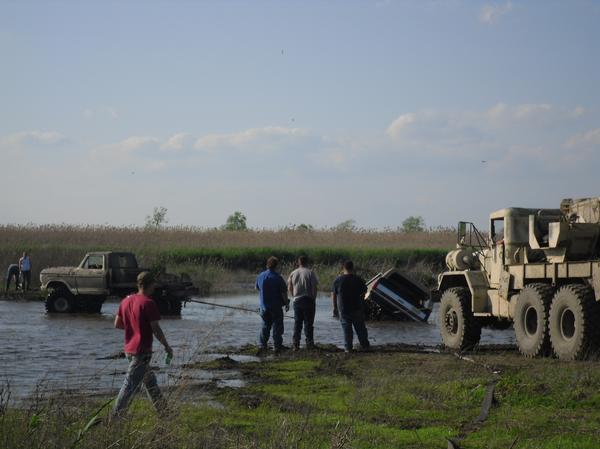 Several people are around a muddy field with water. A tan truck is stuck in the water, and a large military-style truck is nearby