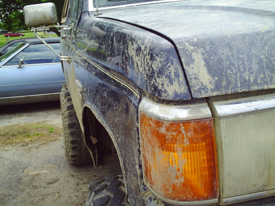 Muddy dark blue truck front fender and headlight. Another car is parked next to it
