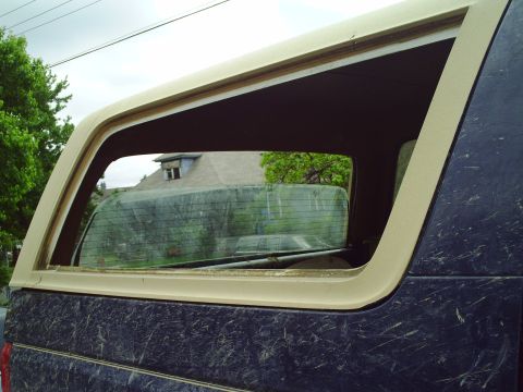 1990 Ford Bronco - The rear side window and dirty windshield of a dark blue SUV with tan trim are visible.