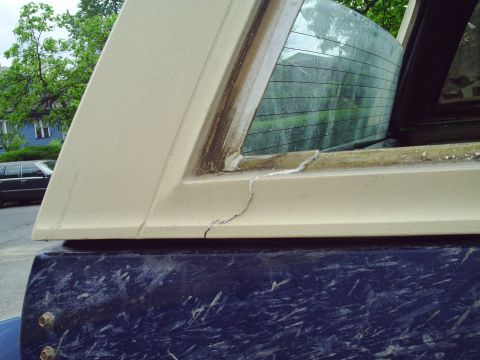 1990 Ford Bronco - Cracked window frame on a vehicle, showing damage to the beige trim and the dark blue body below.
