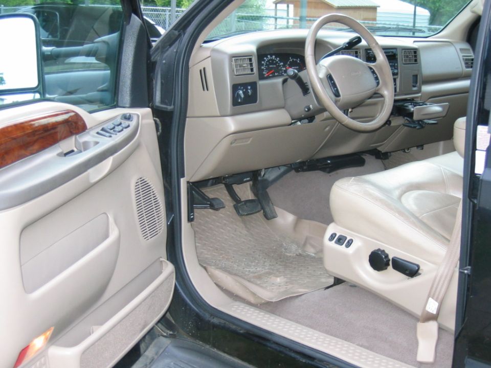 driver's side interior of a truck, showing dashboard, steering wheel, pedals, and tan leather seat