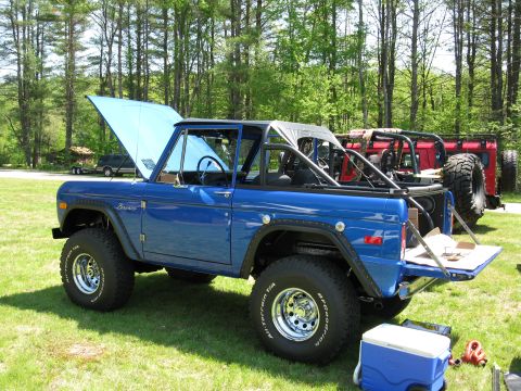 1973 Ford Bronco - A blue 1973 Ford Bronco is displayed with its hood up and tailgate down, parked on a grassy area with trees in the background.