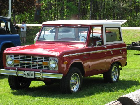 1973 Ford Bronco - A red 1973 Ford Bronco with a white hardtop is parked on a grassy field next to another vehicle.